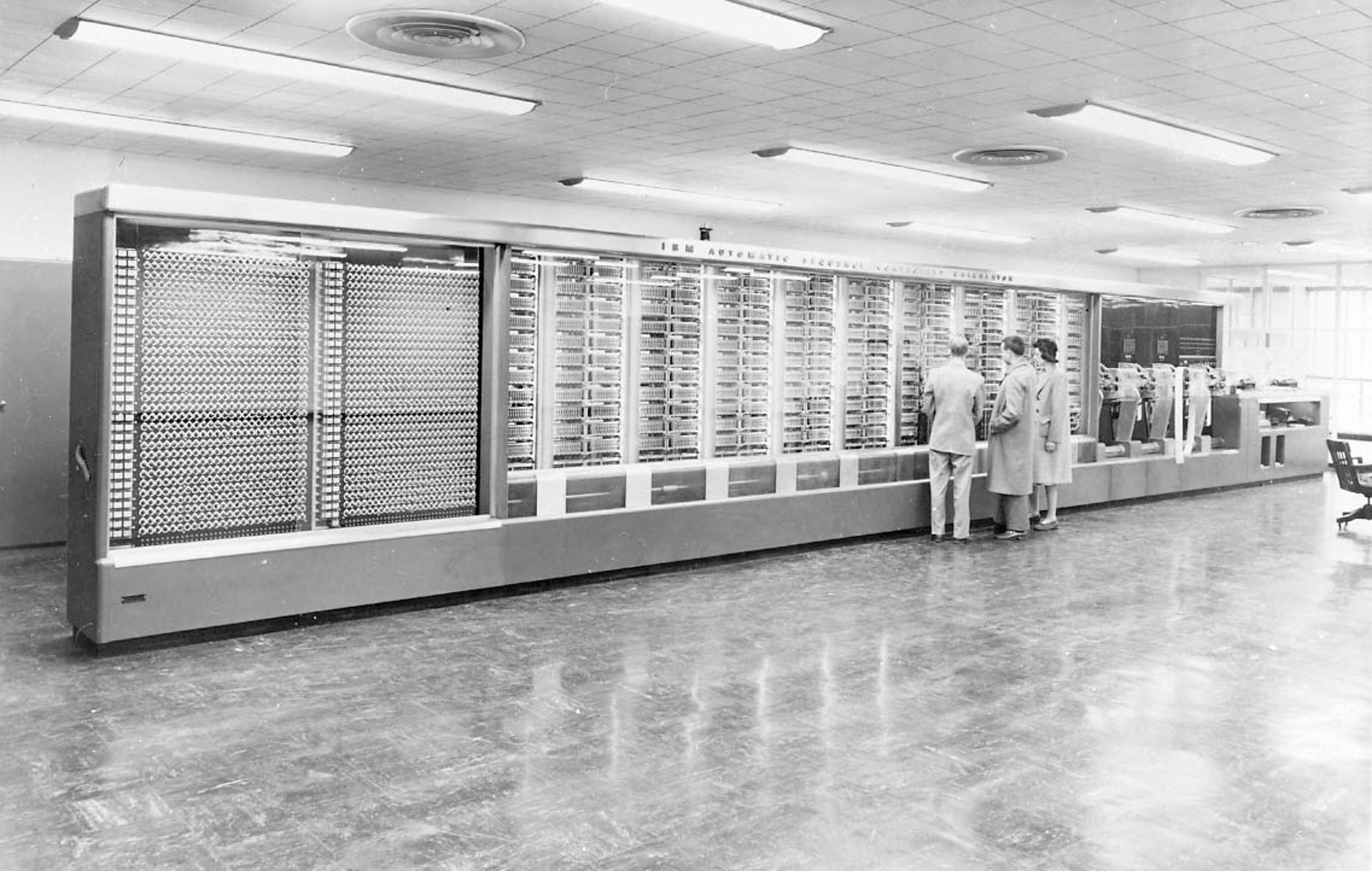Harvard Mark I, an early computer that takes up an entire wall in a large room. There are three people in front of it, investigating missile ballistics data.