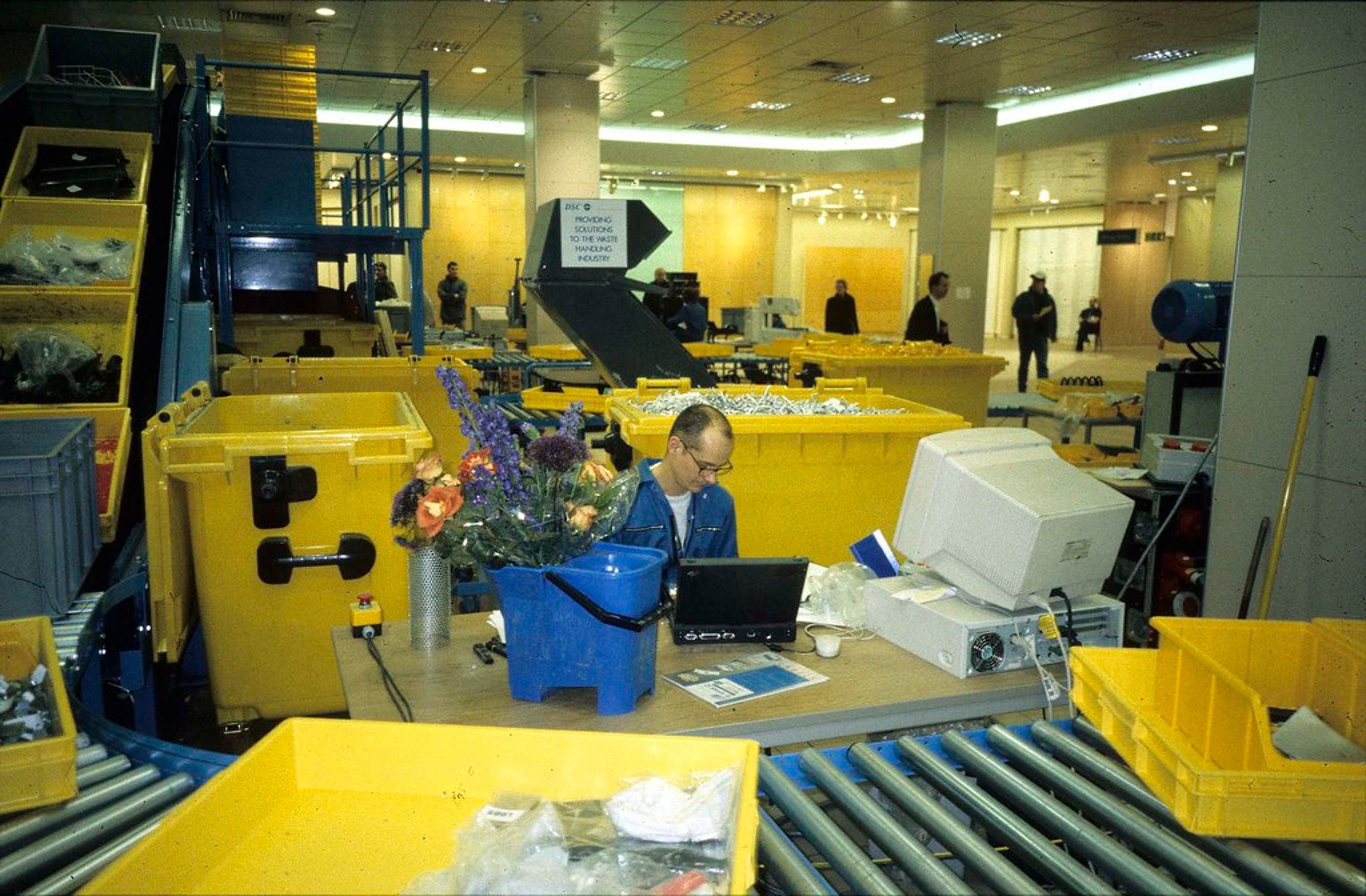 A man in blue industrial suit working behind a desk surrounded by a conveyor system. Boxes and shelves of items are transporting by the conveyor belt.