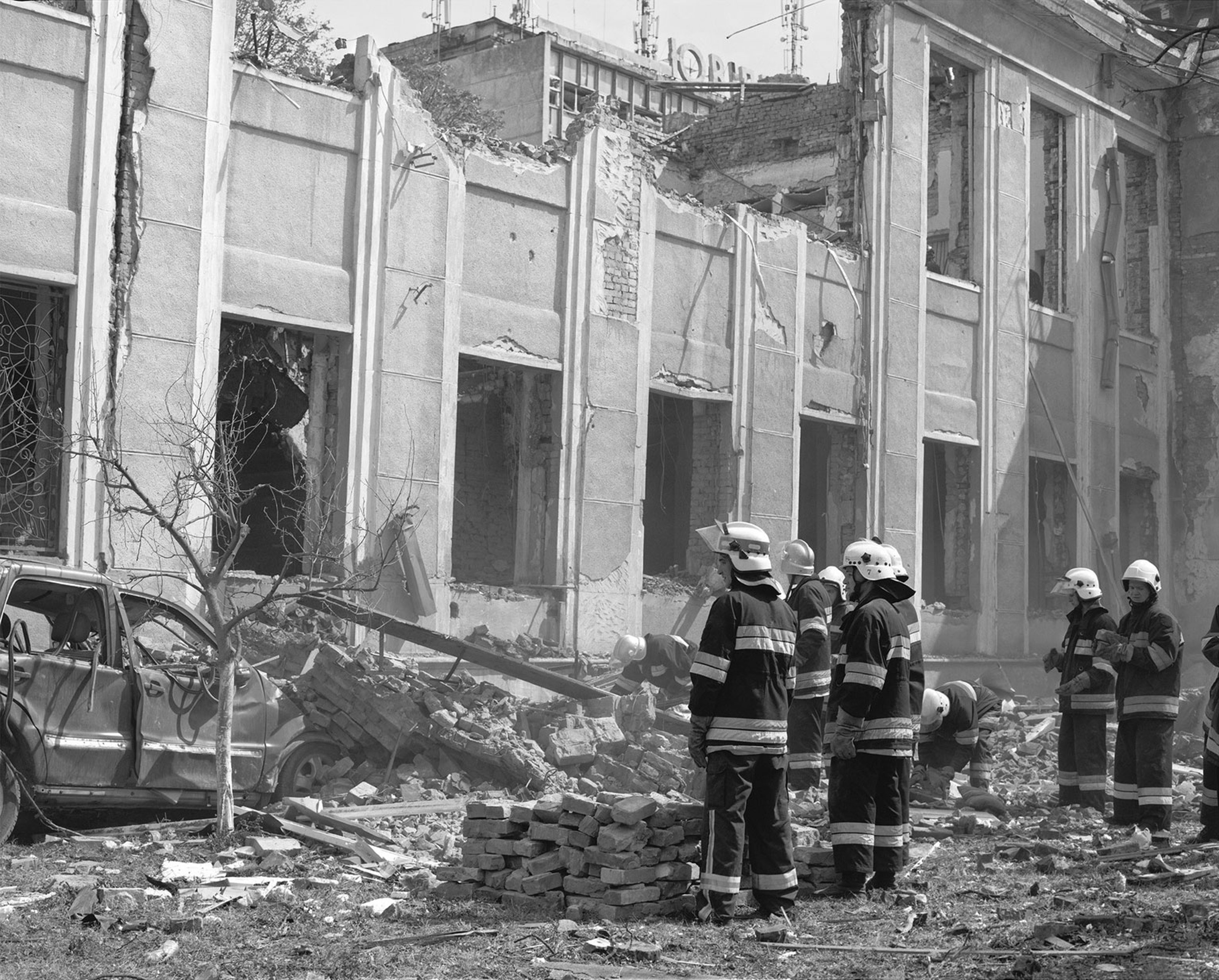 A black and white photography by Yana Kononova. A group of firefighters standing in front of a war-ravaged building.