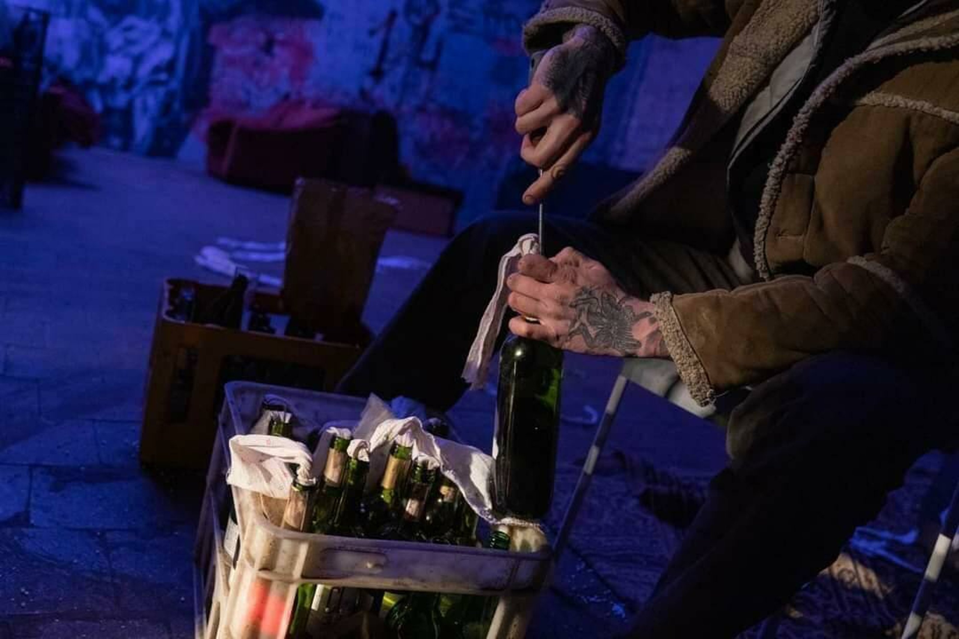 photo from live performance of Otel by Emergency Support Initiative. Two people sitting in a dimly lit room with scribbled-down walls and bottles of alcohol on the floor.