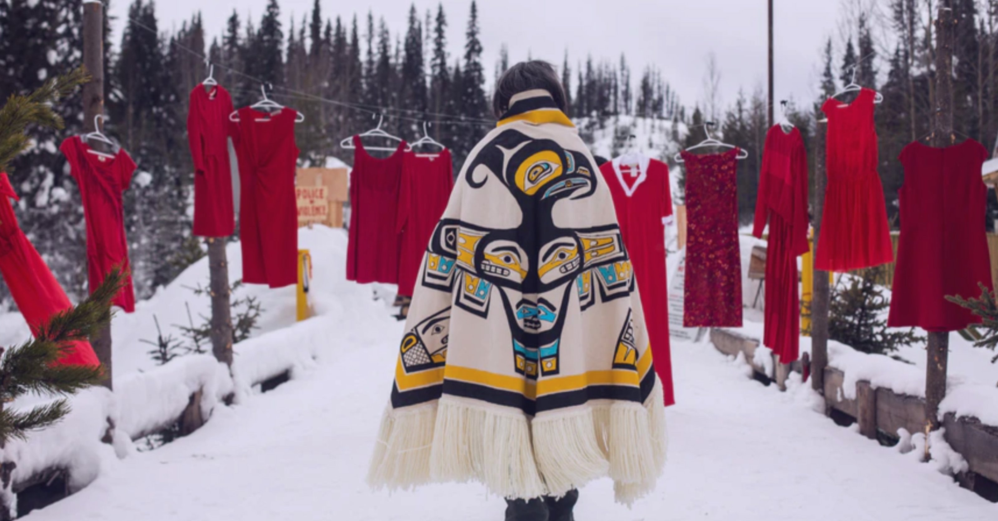 Ts’akë-ze’-Howihkat wearing Native ornamented coat, walking through red dresses hanging in air outdoors