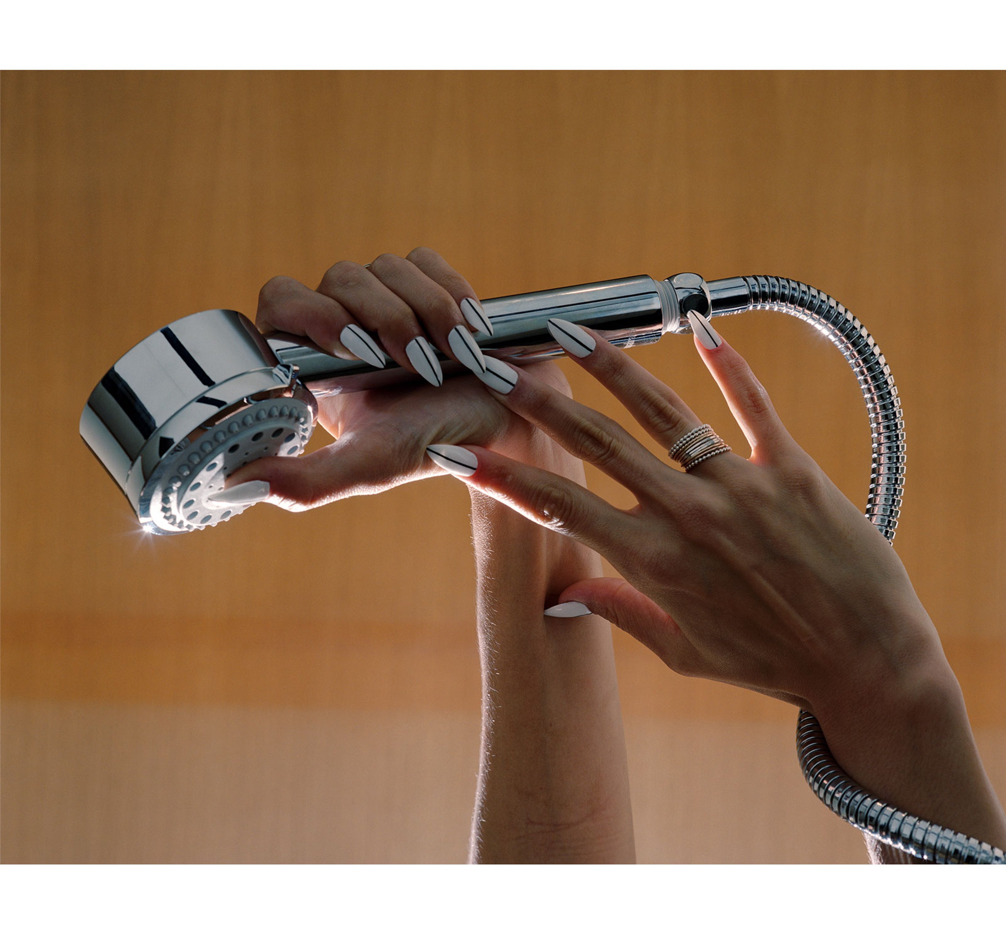 A woman's hands holding up a shower head, the cord wrapped around her arm. She has sharp, painted nails.