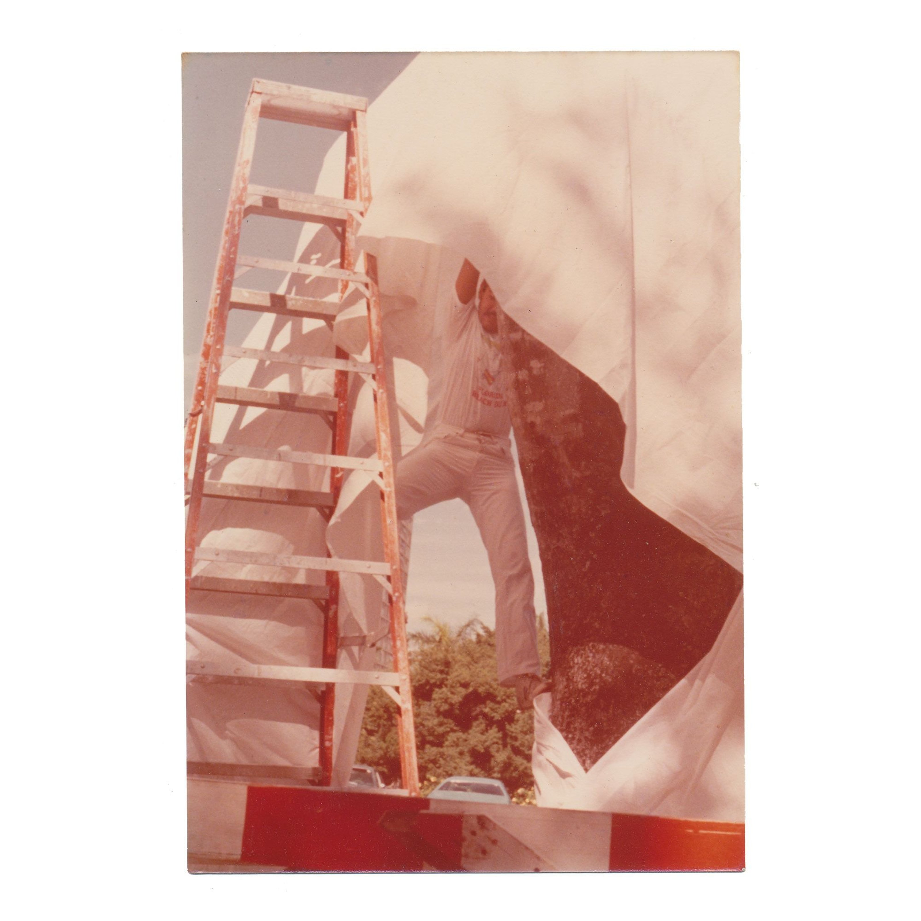 A man installing a sculpture, one foot on a ladder and another lifting off the tarp from a stone structure