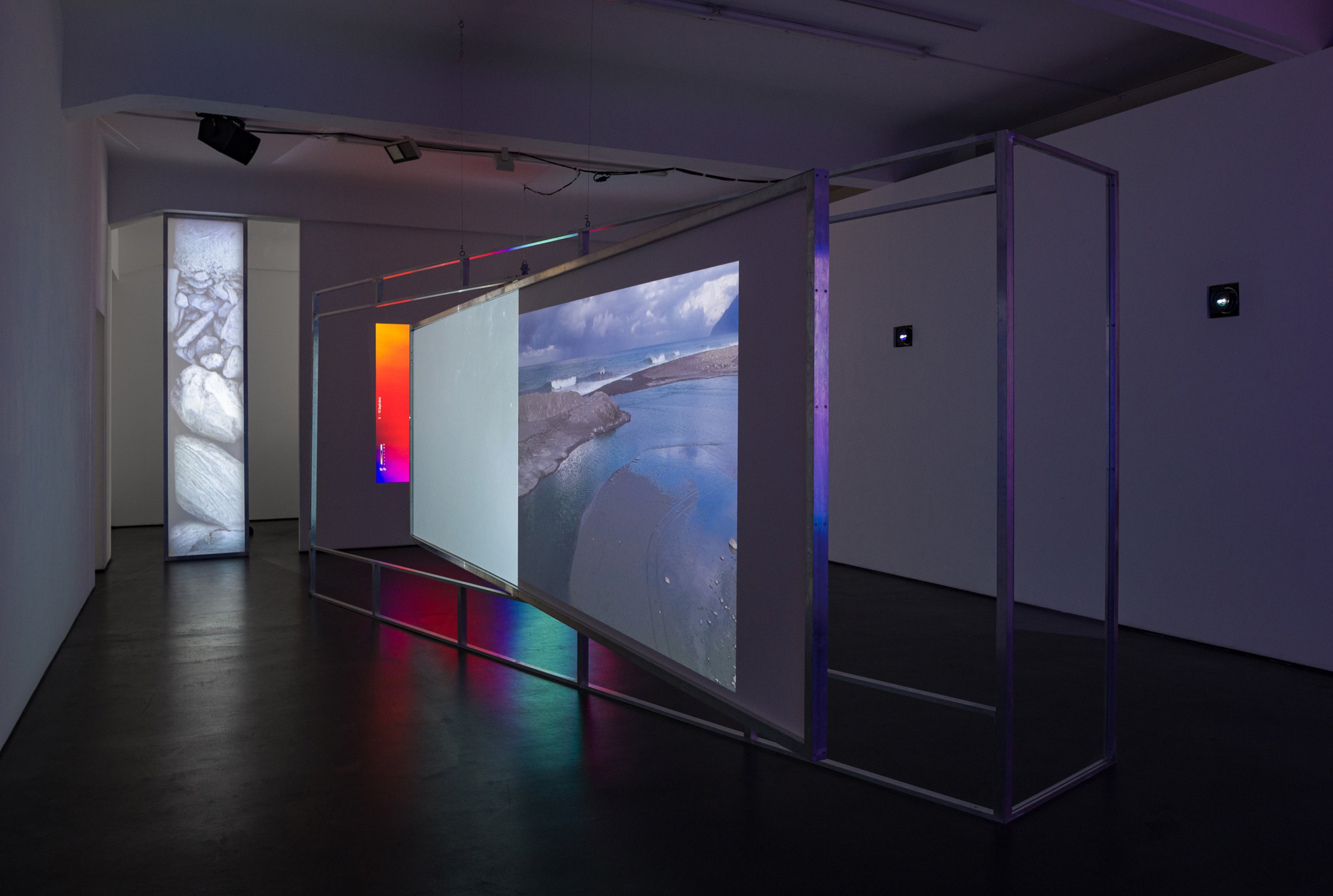 Video installation in gallery, one video is reflected on a screen installed within an aluminum frame, one is vertically placed on a wall, and a third projector screens an image on the wall directly
