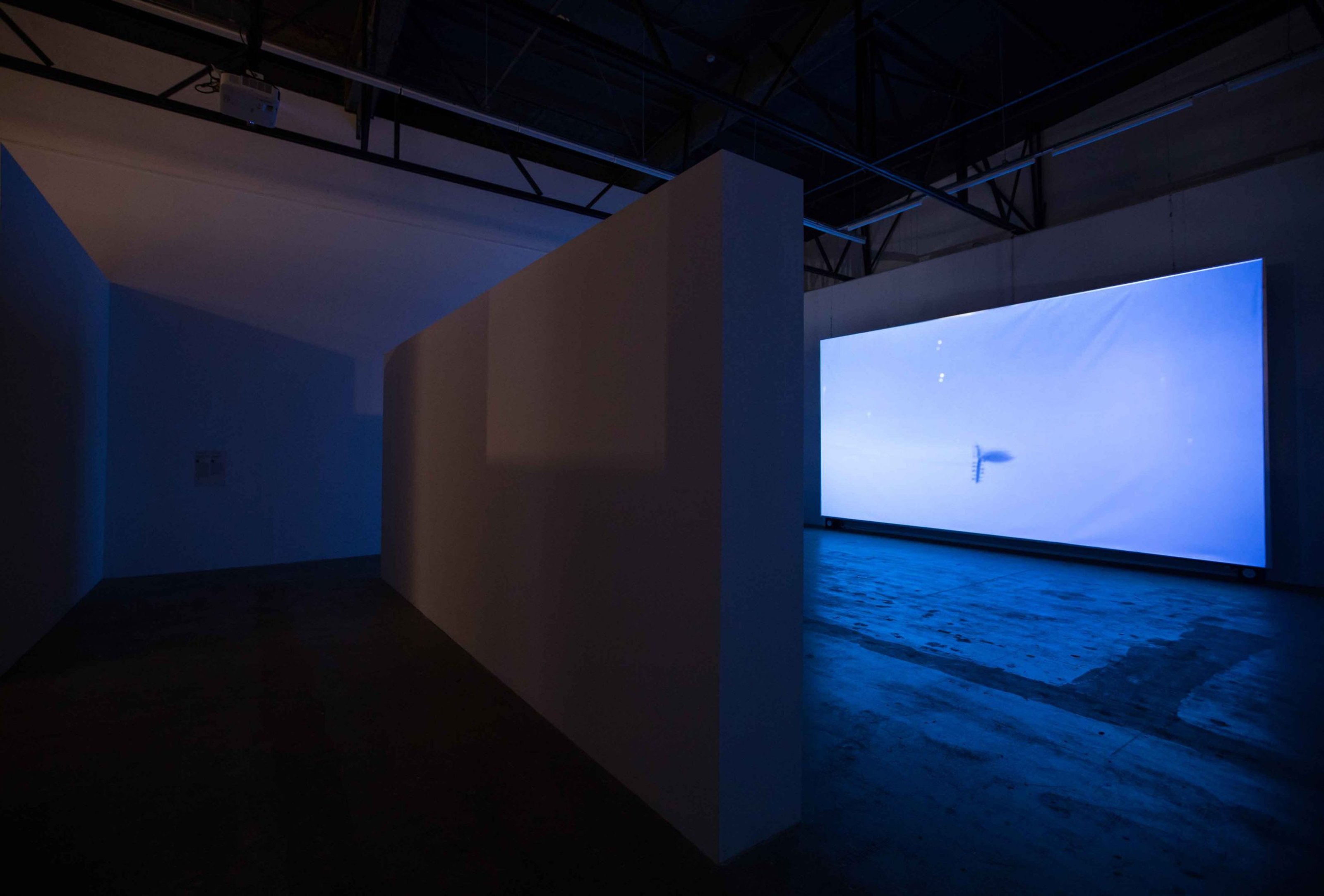 Video reflejado en una pantalla en una pared, mostrando el mar y el cielo y un laúd flotando. Hay otra pared en el centro.