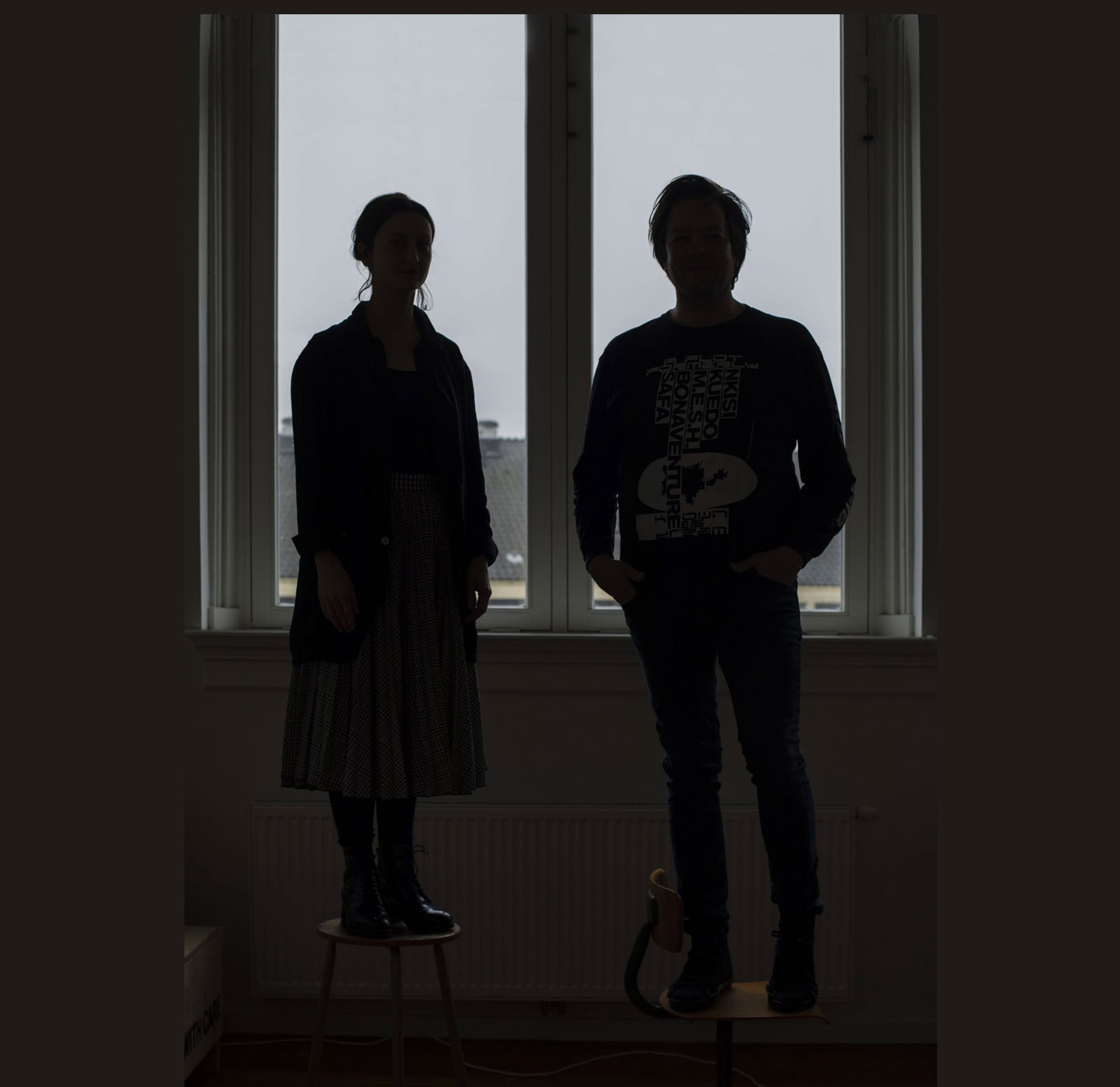Metahaven standing by the window on top of chairs