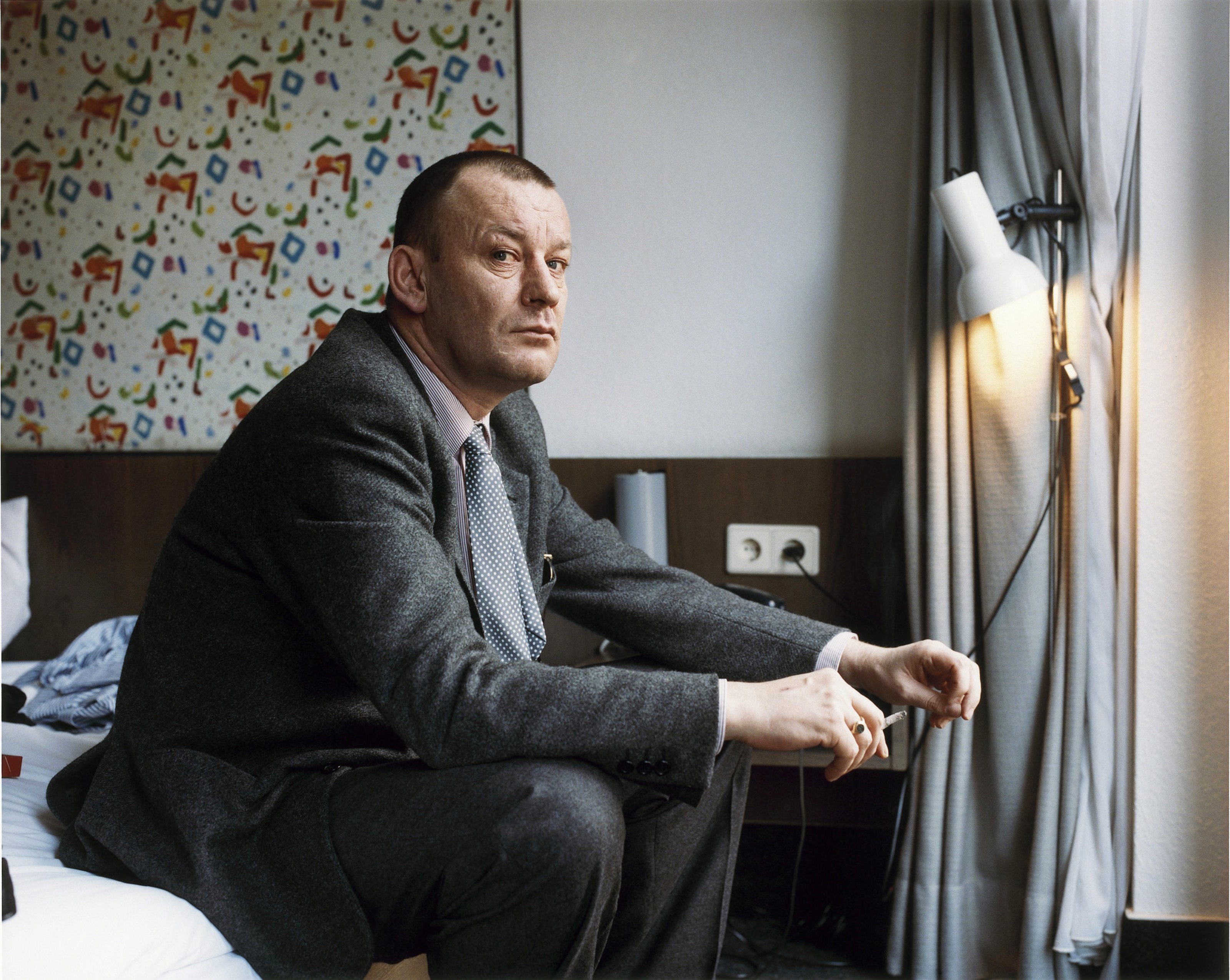 German artist Martin Kippenberger photographed in a suit, smoking while seated on the edge of a bed.